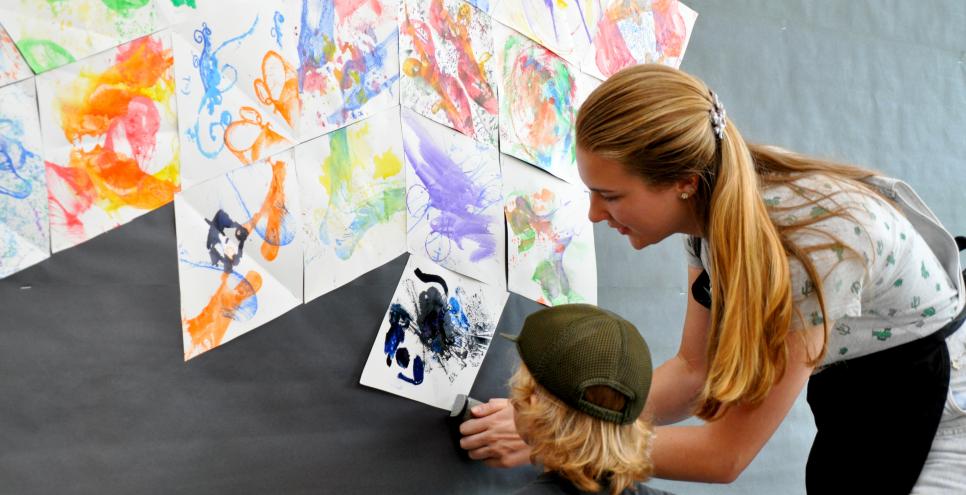 Art Studio intern helps staple a guest's artwork into a wall display as the guest looks on.