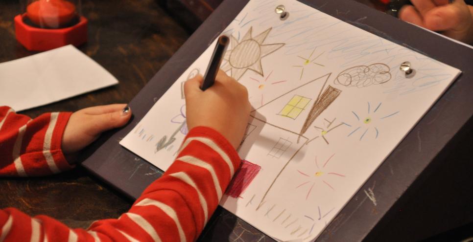 A young child drawing a house and landscape on an angled table.
