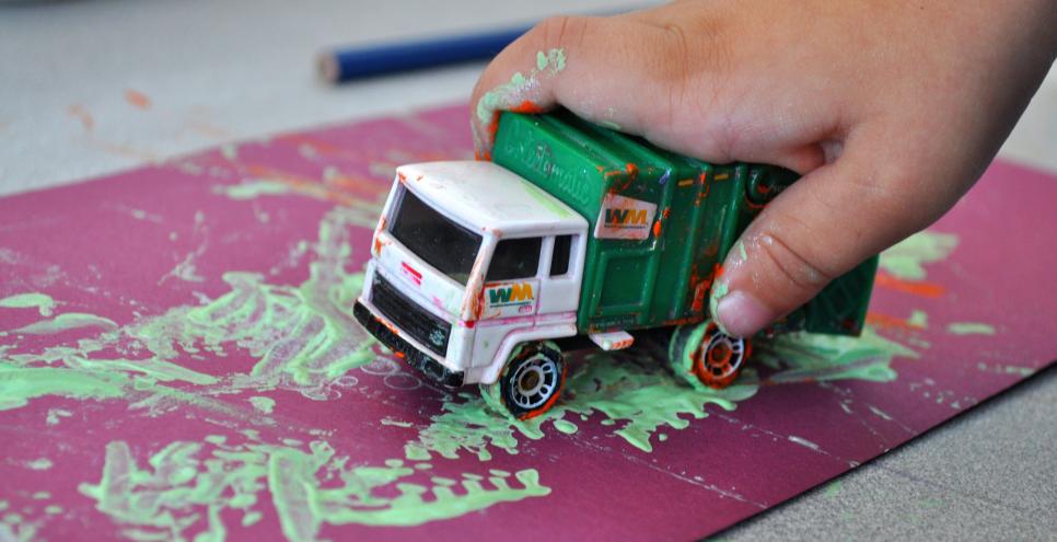 A young child's hand rolling a toy truck through green paint and onto a purple piece of paper.