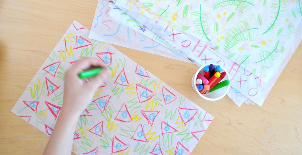 A hand using crayons to draw triangles and shapes onto white tissue paper.
