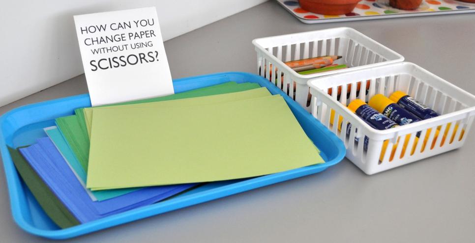 Baskets of drawing tools and glue sticks next to a stack of papers on a tray with a sign that says "how can you change paper without using scissors?"