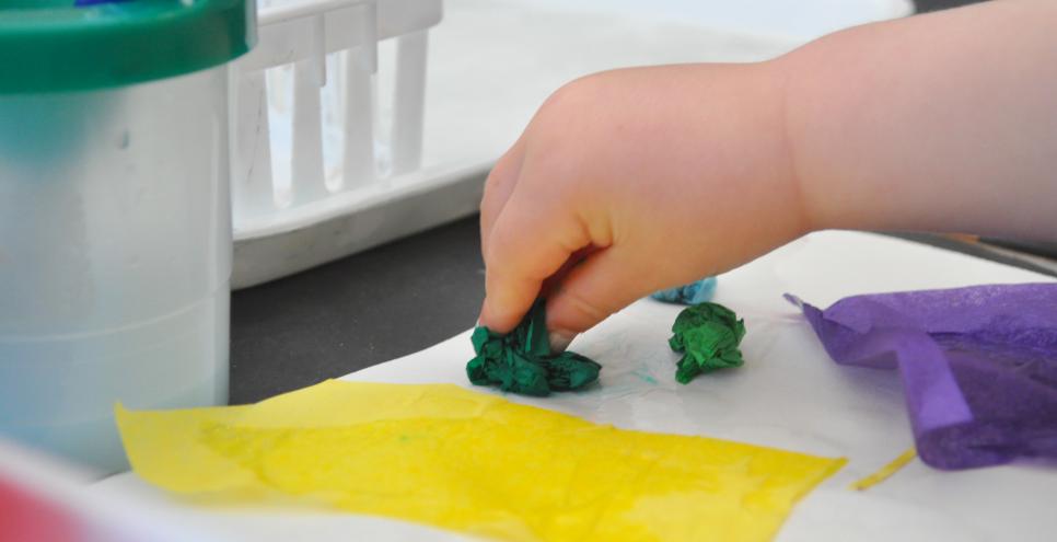 A young child gently places a crumpled piece of green tissue paper onto their background paper with glue.