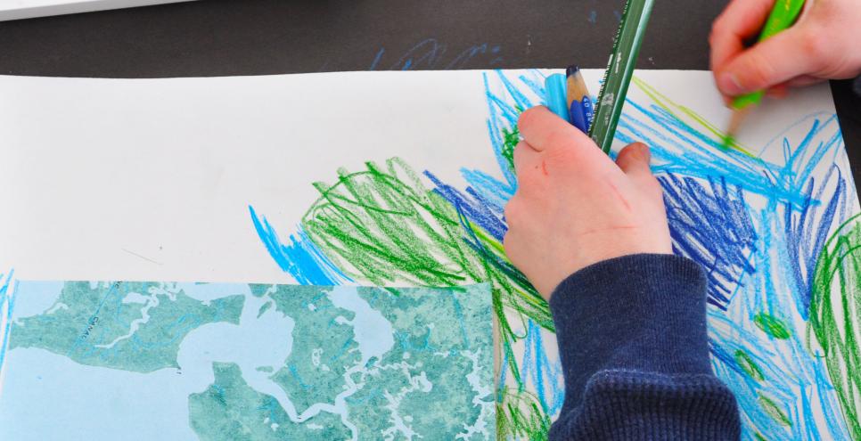 A young child draws with green and blue colored pencils to extend a piece of a map onto a larger piece of white paper.