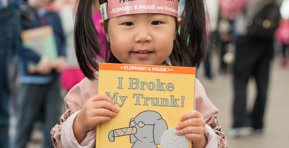 Child wearing pink Piggie ears and holding Elephant & Piggie book.