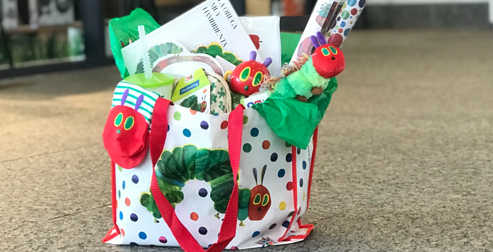 A Very Hungry Caterpillar tote bag filled with Caterpillar products is set on the floor in front of the museum shop.