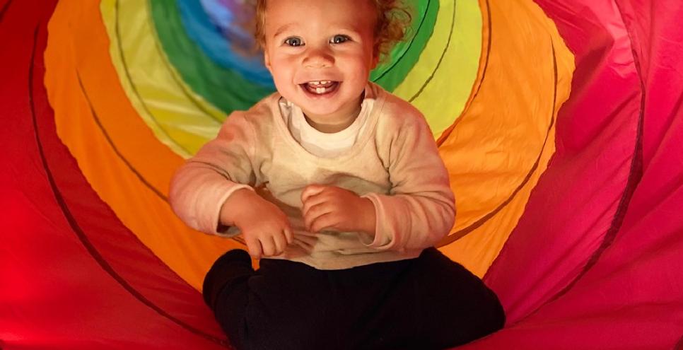 Smiling child sits inside tunnel of rainbow colors.