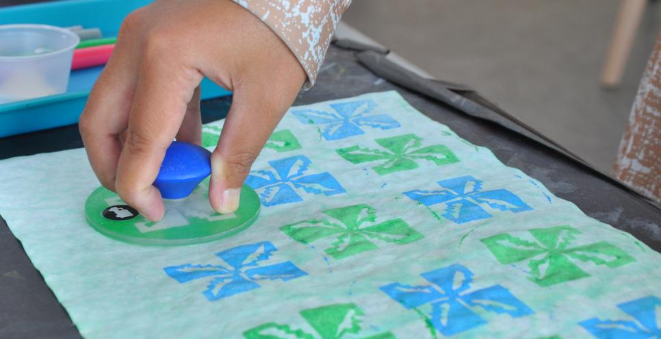A hand presses a painted stamp onto a piece of dyed paper, creating a pattern inspired by batik.
