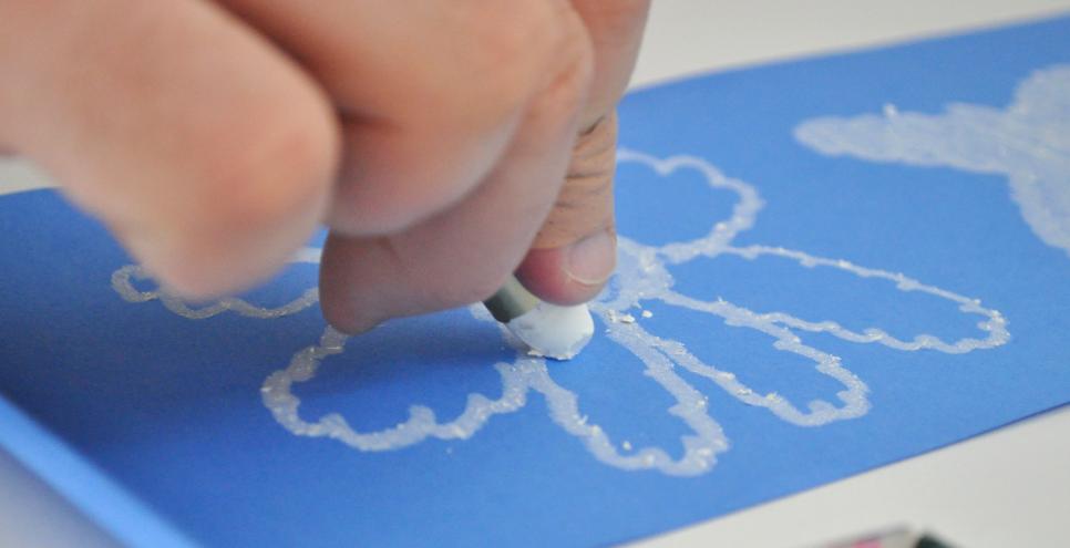 A hand draws a flower using a white oil pastel on blue paper.