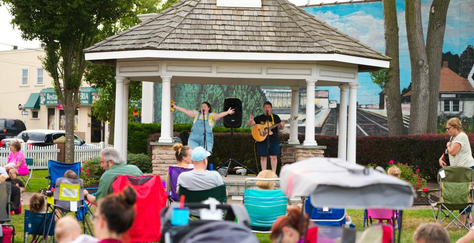 Two people under gazebo playing singing and playing instruments to a crowd.