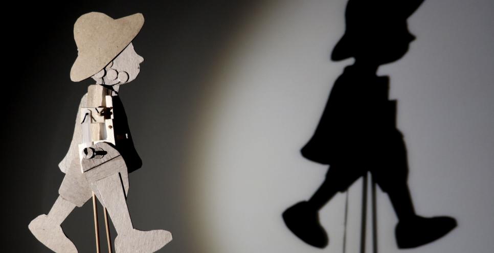 A puppet of a man in a hat walking, with the puppet's shadow cast on a wall.