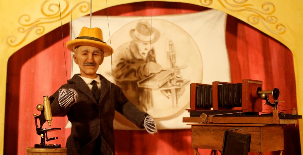 Marionette puppet of a man in a suit and hat next to a microscope and camera.