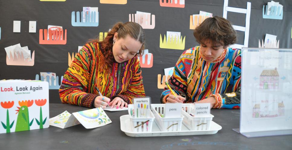 Two people sitting at a table drawing, art materials and books also on the table