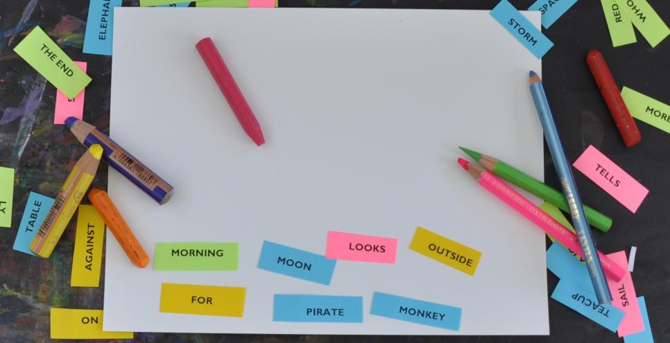 A blank piece of paper on a black background. There is an array of materials, pencils, crayons and watercolor pencils scattered across the table top. There are colorful mini strips of paper that have one word on them each. Someone has taken the words and rearranged them on the blank page saying "Morning moon looks outside for pirate monkey"