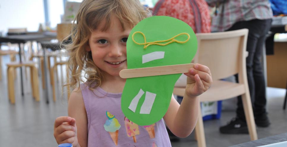 A child excitedly showing their artwork. Holding up a collaged paper with yarn, papers and a popsicle stick.