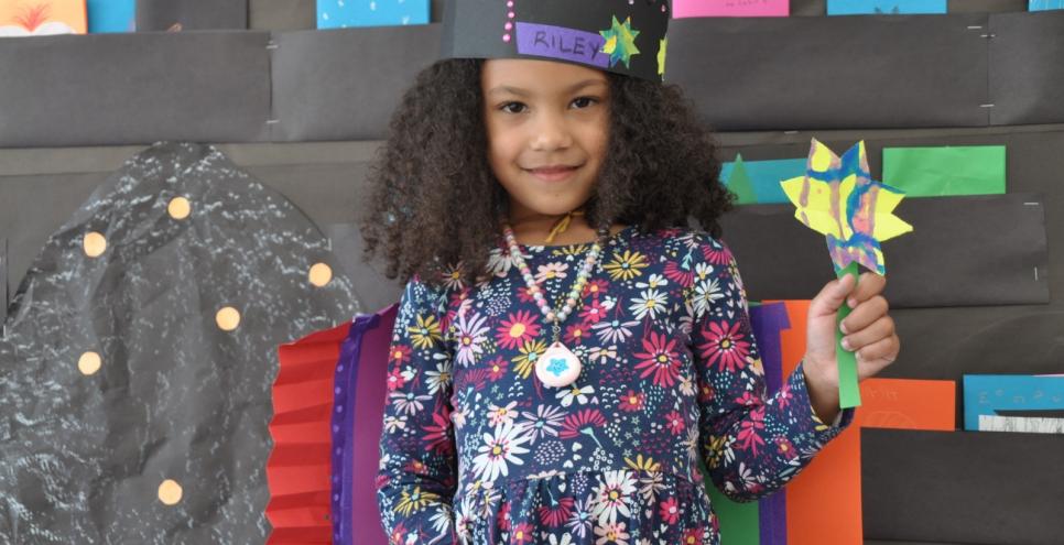 A young artist shows off their paper creations. A bright star shaped wand is held up with pride, a rainbow paper cape rests on the shoulders and a star crown sits on top of the head. 