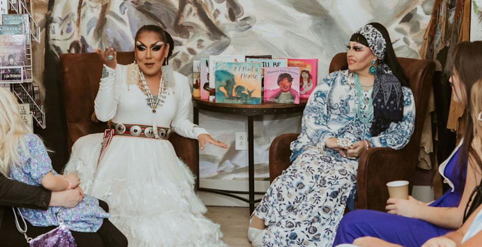 Two drag storytellers sit next to a display of picture books for storytime.