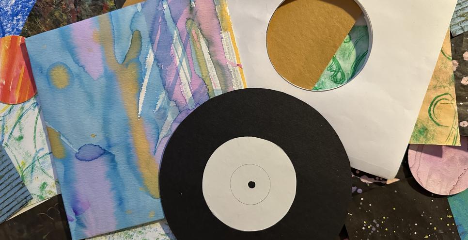 A variety of colorful collage papers in the background with a vinyl record cut out of black card stock laying on top of a blank white record inner sleeve and a colorful record jacket with light blue, pink, and peach paint. 