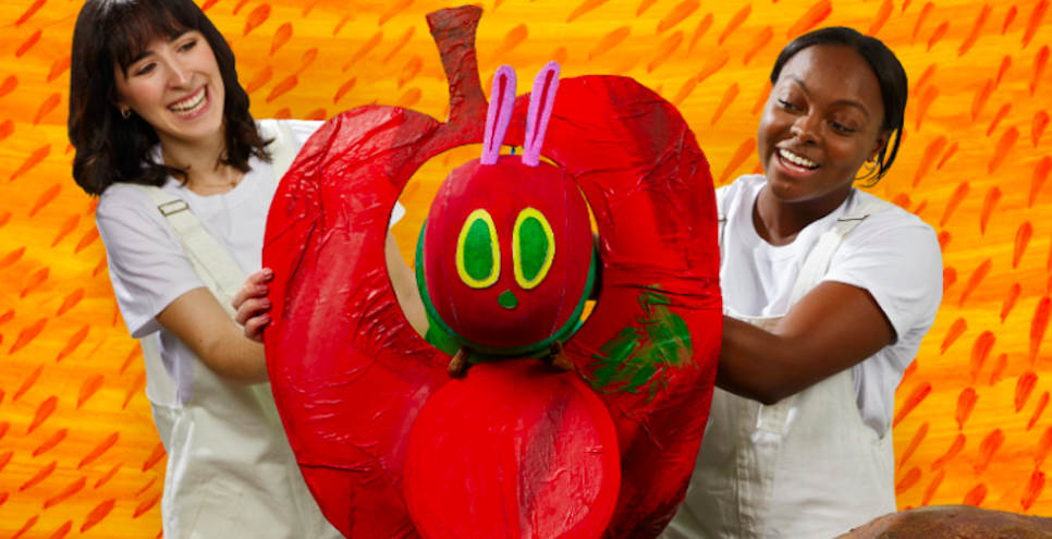 Two puppeteers smiling with a Very Hungry Caterpillar puppet eating through a large red apple.