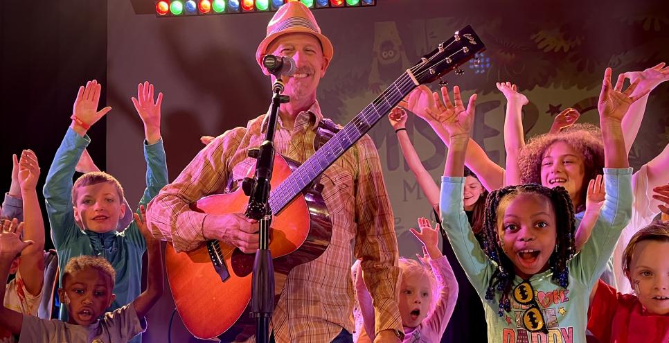 Mister G holding a guitar surrounded by children with their hands raised in the air.