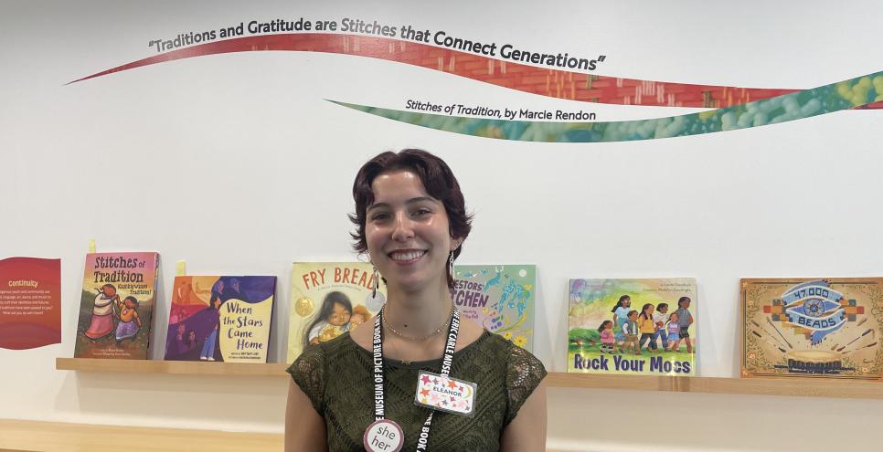 Young woman with short brown hair with a Museum lanyard around her neck and picture book exhibition in the background 