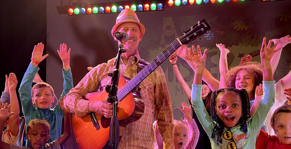 MISTER G standing in front of a microphone holding a guitar surrounded by smiling children with their hands up.