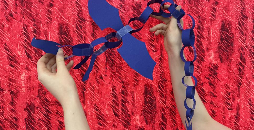 A blue paper chain dragon against a red fabric background. hands holding the paper creation up. 