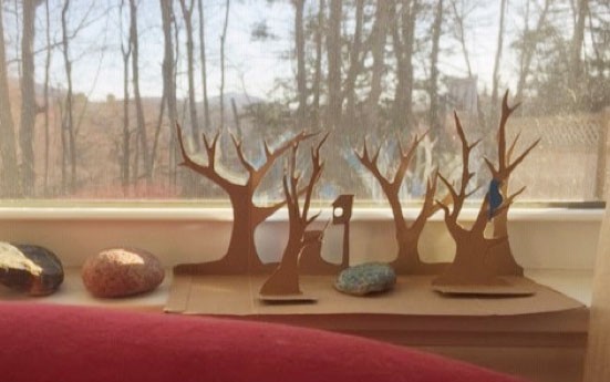 Cardboard tree silhouettes cut out of a box sit in front of a window with a view of hills, trees, and sky. 