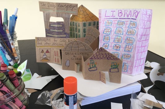 A cardboard city made of cardboard folded to stand up. Buildings are colored in with windows, doors and architectural details. Some are labelled with the building's function such as "Library" and "Observatory for Science".