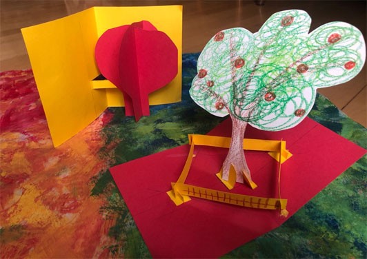 Two accordion fold pop-ups, the yellow card with the red tree, and a red card with a yellow fence and a tree that has a brown trunk, green canopy, and red fruit. The red card has the tree in the middle and the fence surrounds the tree.