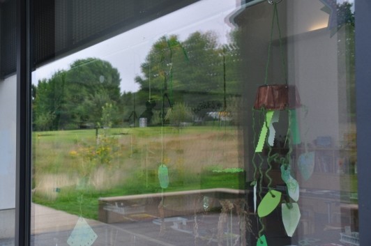 A plant mobile in the window with a reflection of Bobbie’s Meadow on the left.